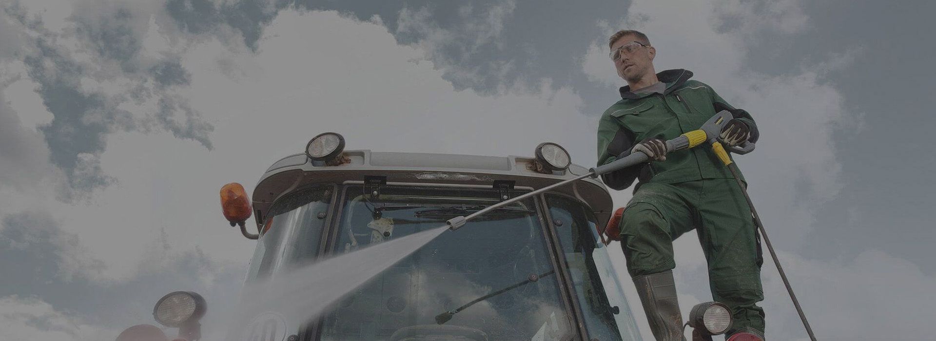 Eine Person in grüner Arbeitskleidung reinigt bei bewölktem Himmel einen Traktor mit einem Hochdruckreiniger und demonstriert damit die Wartung in der Landwirtschaft.