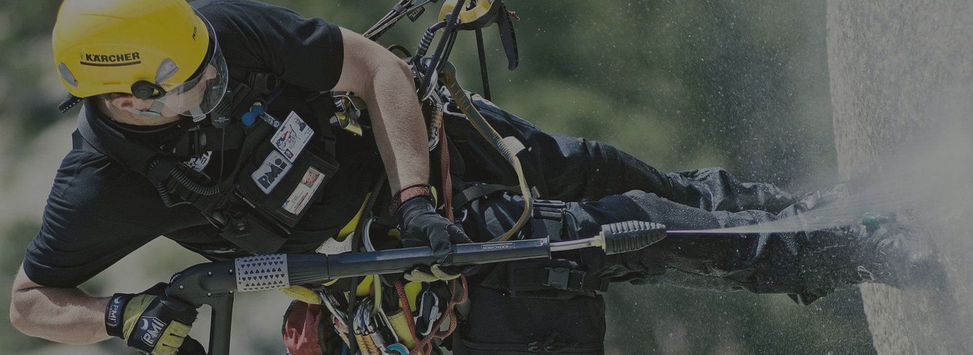 Ein Arbeiter mit gelbem Helm reinigt mit einem Hochdruckreiniger eine Felsoberfläche, während er an einem Sicherheitsgeschirr hängt.